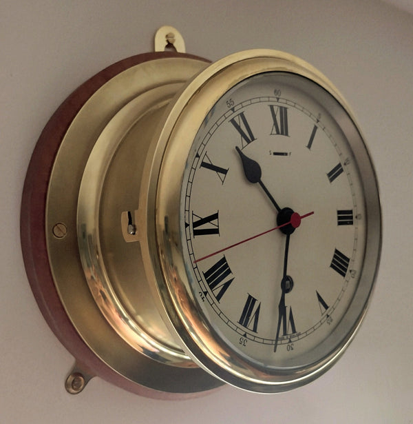 A classic nautical-style wall clock with a polished brass case, Roman numeral dial, and a mahogany backing plate. Its ship’s porthole design gives it a timeless maritime look, making it a striking decorative piece.