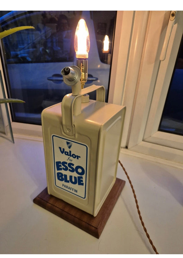 A vintage-style table lamp made from a retro “Valor for Esso Blue Paraffin” fuel can, mounted on a mahogany base. The flame-shaped bulb and classic branding give it a warm glow and a distinctive industrial, nostalgic look.